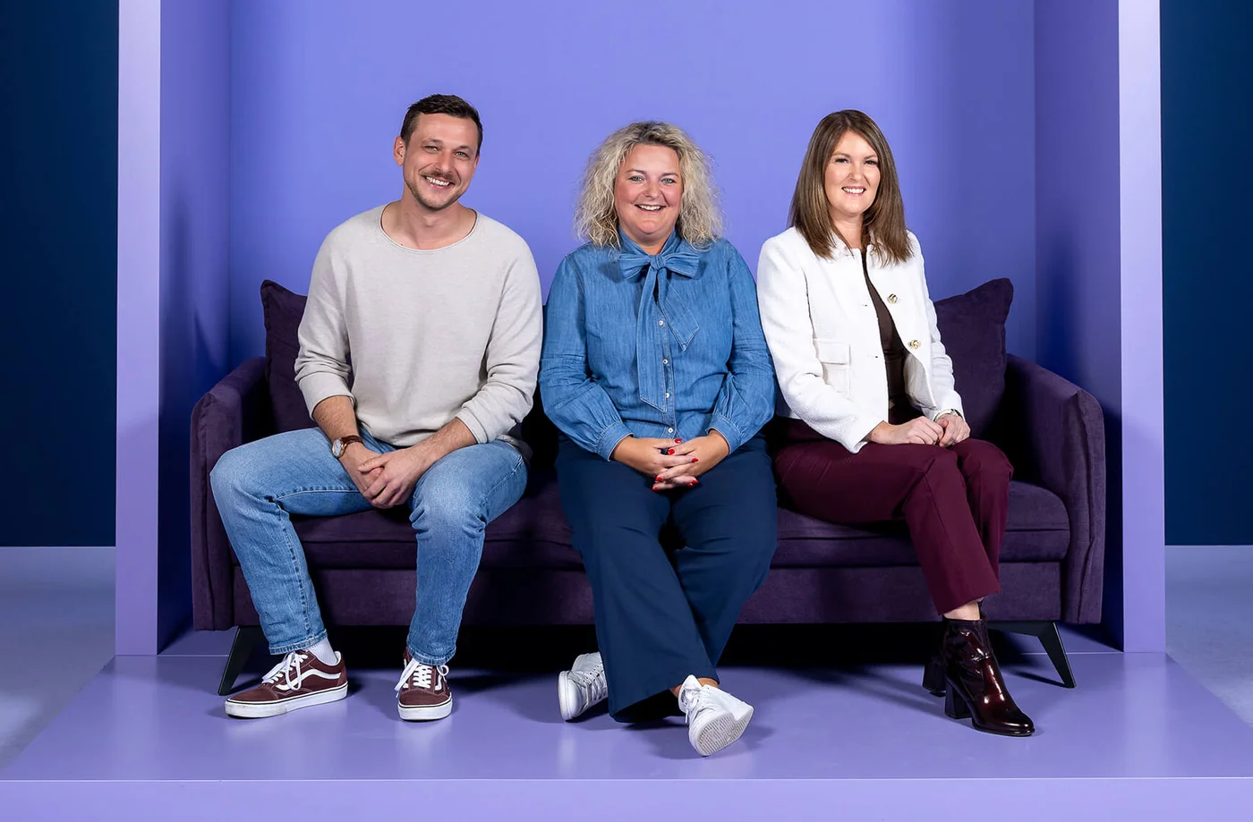 Ein Mann und zwei Frauen sitzen auf einem lila Sofa vor einem Hintergrund mit der Aufschrift "INSIDE". Alle lächeln in die Kamera.