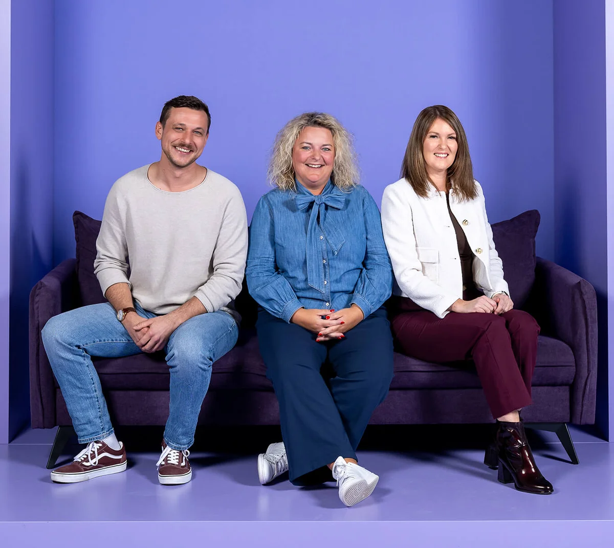 Ein Mann und zwei Frauen sitzen auf einem lila Sofa vor einem Hintergrund mit der Aufschrift "INSIDE". Alle lächeln in die Kamera.