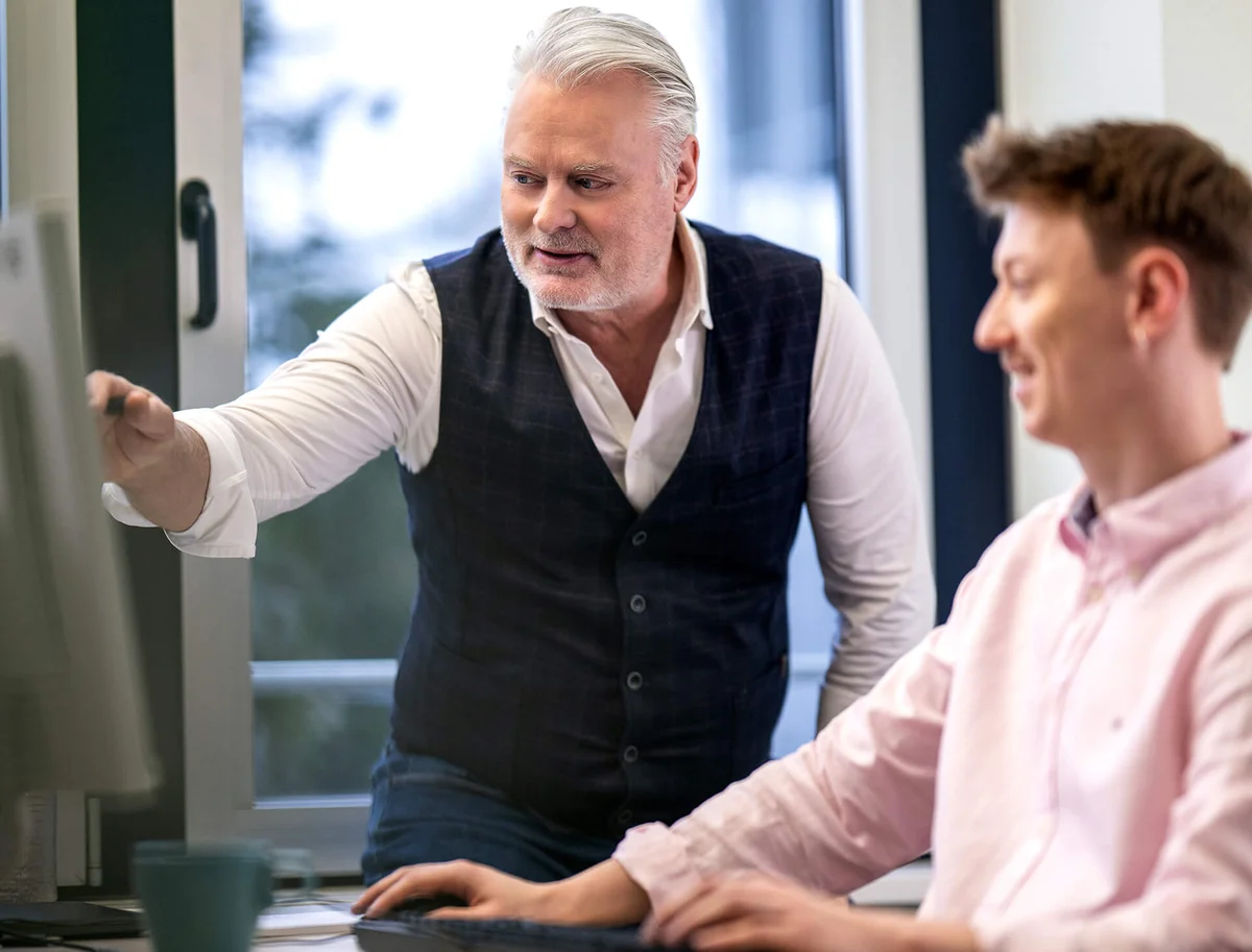 Ein älterer Mann zeigt einem jüngeren Mann an einem Computerbildschirm etwas. Der jüngere Mann sitzt und arbeitet am Computer, während der ältere Mann steht und auf den Bildschirm zeigt. Sie scheinen in einem Büro zu sein.