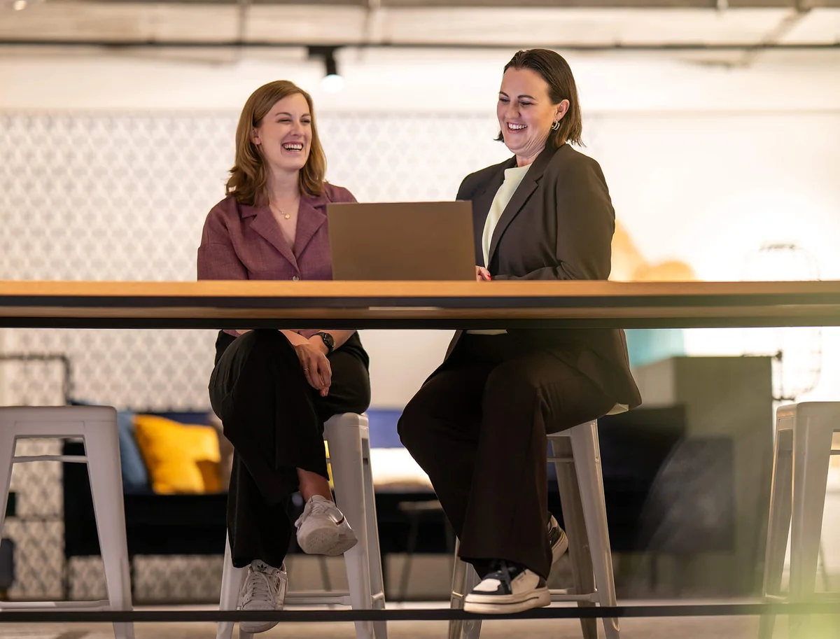 Zwei lachende Frauen sitzen auf Barhockern an einem Tisch, während sie auf einen Laptop schauen. Eine trägt einen Blazer, die andere ein lila Oberteil. Der Hintergrund ist unscharf mit moderner Dekoration.