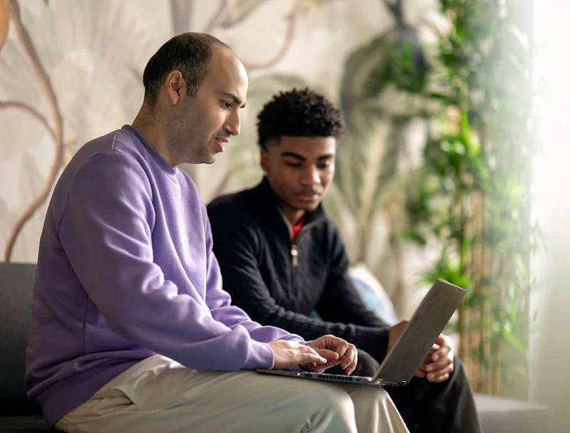 Zwei Männer sitzen auf einem Sofa, der eine im lila Pullover tippt auf einem Laptop, während der andere im schwarzen Pullover zuhört. Sie scheinen konzentriert zu sein und in einem Gespräch oder einer Zusammenarbeit vertieft.
