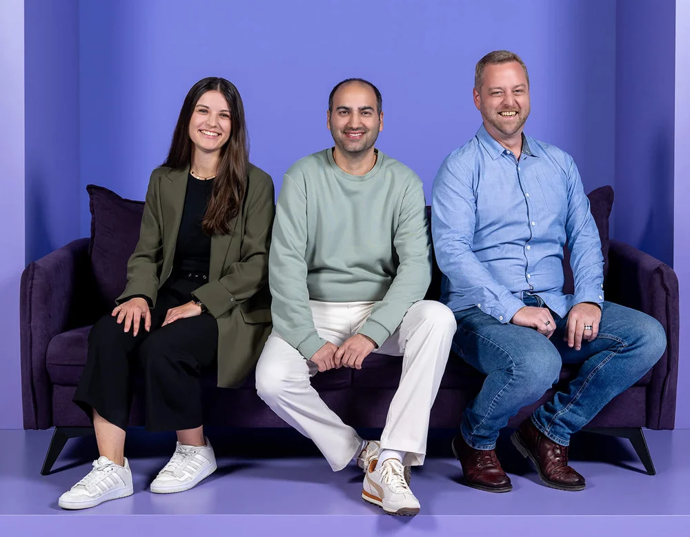 Drei Personen sitzen auf einem Sofa vor einem lila Hintergrund. Hinter ihnen steht in großen Buchstaben der Schriftzug "INSIDE". Alle drei lächeln und schauen in die Kamera.