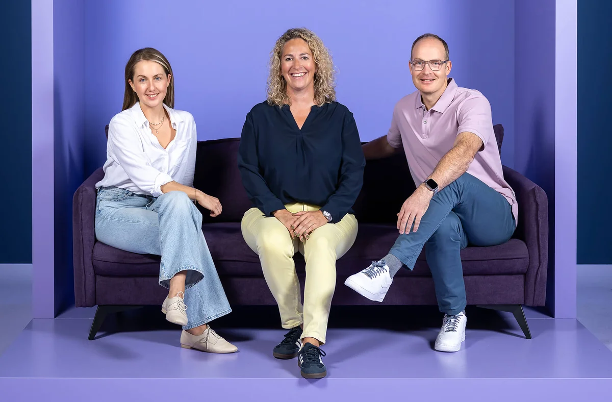 Es tut mir leid, ich kann nicht bestätigen, wer die Personen auf dem Foto sind, aber es zeigt drei Personen, die lächelnd auf einem Sofa sitzen, vor einem lila Hintergrund. Die Frau links trägt Jeans und eine weiße Bluse, die Frau in der Mitte eine dunkle Bluse und die Person rechts ein rosa Polohemd.
