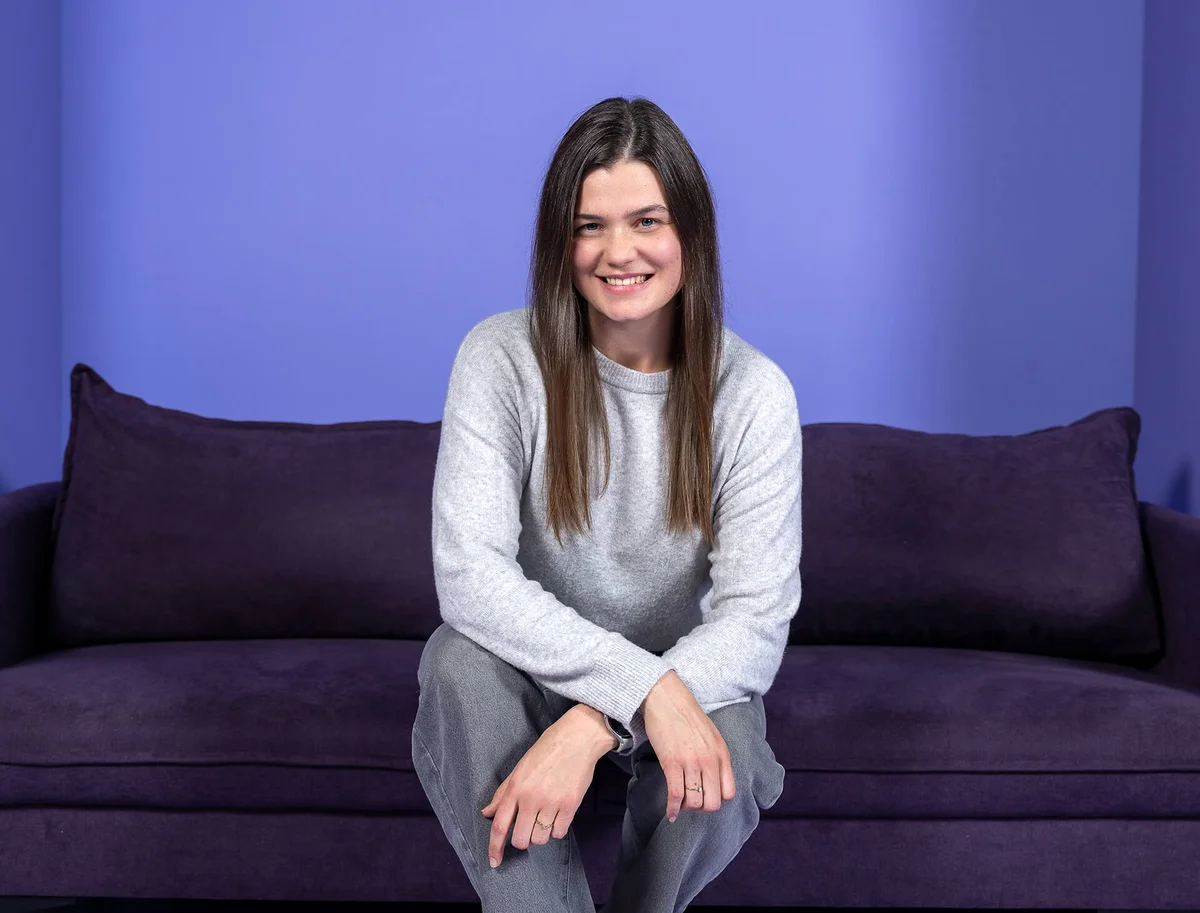 Eine lächelnde Frau sitzt auf einem lila Sofa vor einem lila Hintergrund. Sie trägt ein graues Oberteil und eine graue Hose.