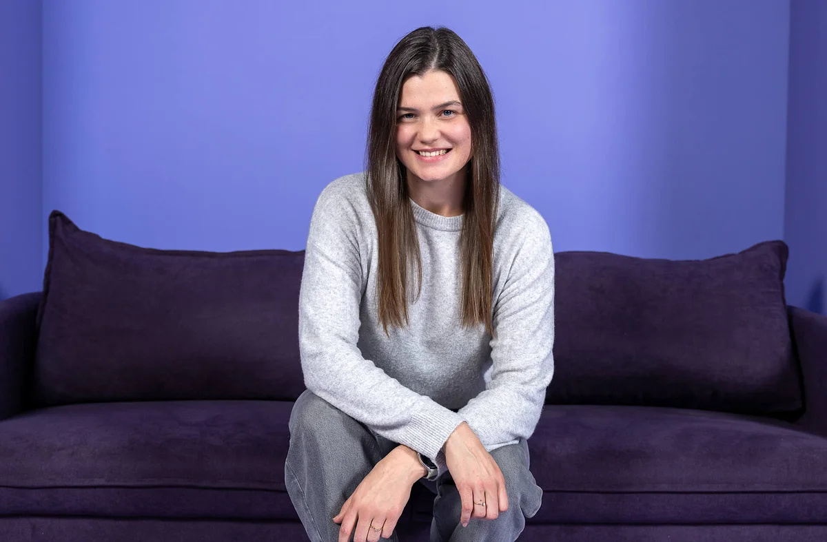 Eine lächelnde Frau sitzt auf einem lila Sofa vor einem lila Hintergrund. Sie trägt ein graues Oberteil und eine graue Hose.