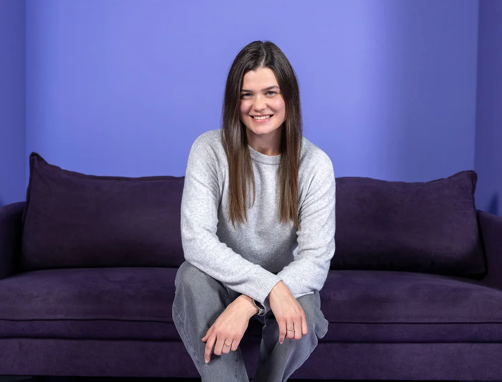 Eine lächelnde Frau sitzt auf einem lila Sofa vor einem lila Hintergrund. Sie trägt ein graues Oberteil und eine graue Hose.