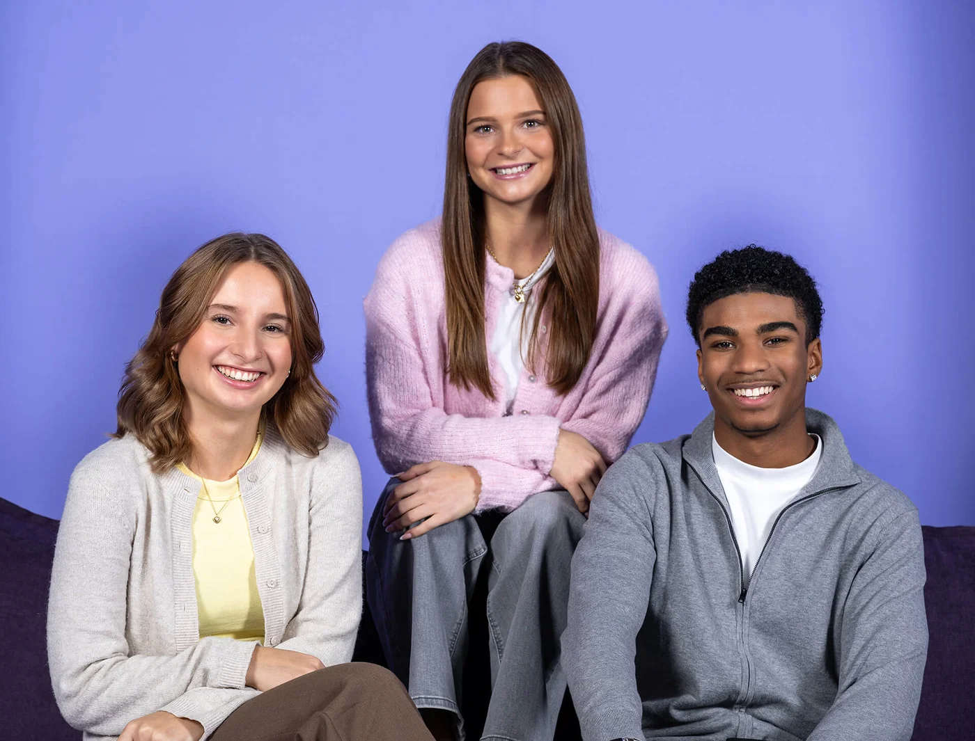 Drei lächelnde junge Erwachsene sitzen vor einem violetten Hintergrund. Zwei Frauen und ein Mann tragen legere Kleidung in hellen Farben. Sie schauen freundlich in die Kamera.