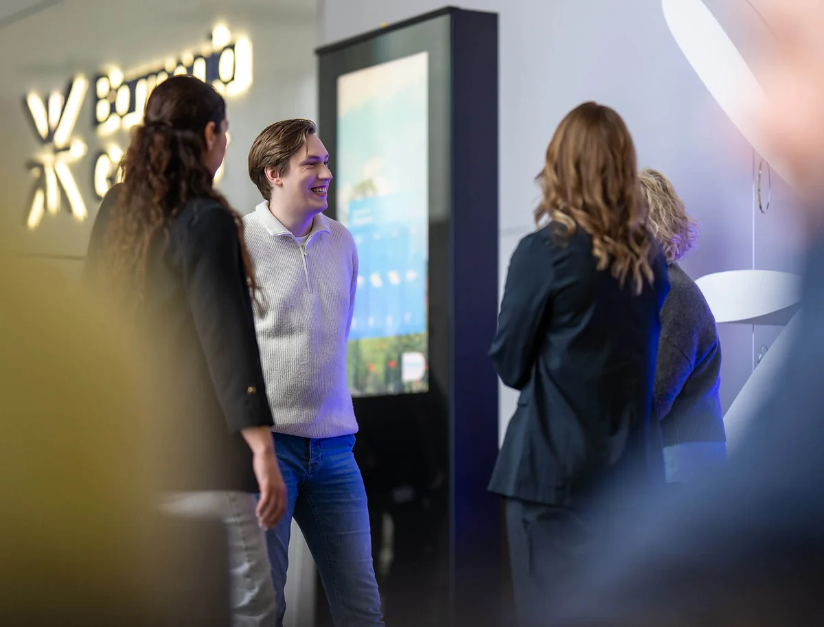 Eine Gruppe von vier Personen steht in einem freundlichen Gespräch vor einem digitalen Bildschirm. Eine Person trägt einen weißen Pullover und die anderen formelle Kleidung. Sie wirken entspannt und lächelnd. Im Hintergrund ist ein beleuchtetes Logo sichtbar.