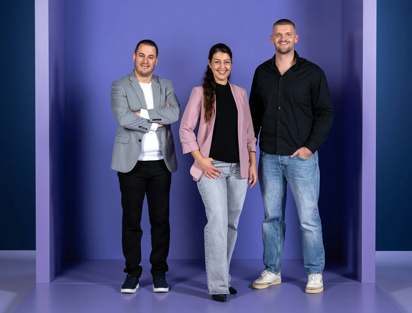 Drei fröhliche Personen stehen nebeneinander vor einem lilafarbenen Hintergrund. Zwei Männer und eine Frau tragen legere Kleidung. Der Mann links trägt einen grauen Blazer, die Frau in der Mitte ein rosa Jackett, und der Mann rechts ein schwarzes Hemd. Alle lächeln in die Kamera.