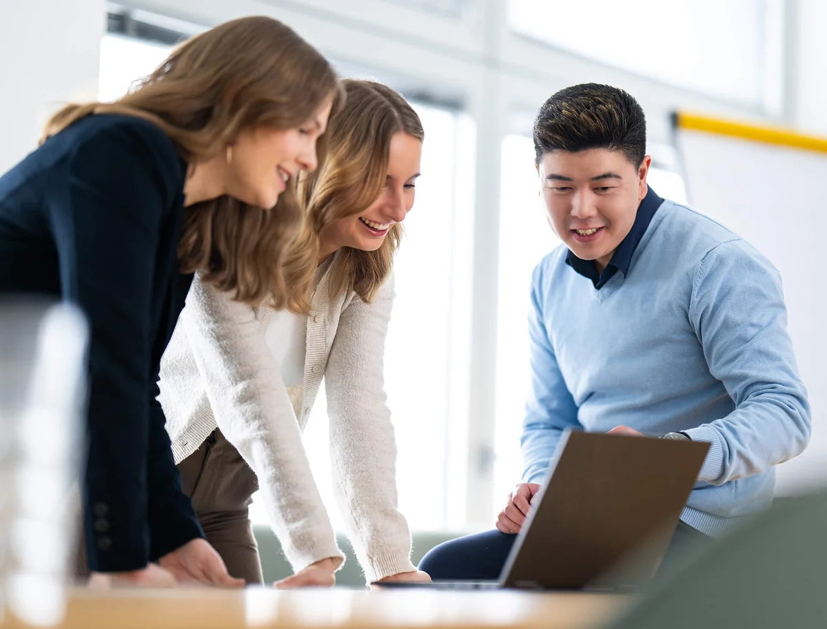 Drei Personen stehen um einen Laptop und blicken lachend auf den Bildschirm. Sie sind in einem modernen Büro und wirken konzentriert und engagiert. Die Gruppe scheint konstruktiv zu arbeiten oder an einer Präsentation teilzunehmen.