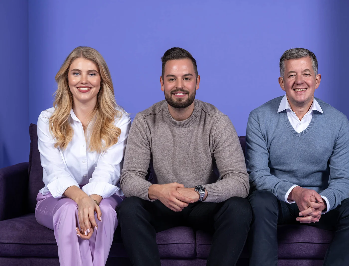 Drei Personen sitzen auf einem Sofa vor einem lilafarbenen Hintergrund. Die Frau links trägt eine weiße Bluse, der Mann in der Mitte einen grauen Pullover, der Mann rechts ein blaues Oberteil. Alle lächeln und schauen in die Kamera.