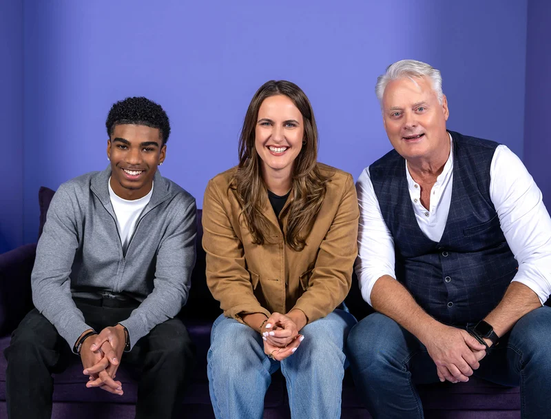 Drei Personen sitzen auf einem Sofa vor einem lila Hintergrund. Eine Frau sitzt in der Mitte zwischen zwei Männern. Alle lächeln freundlich in die Kamera. Die Szene wirkt entspannt und herzlich.