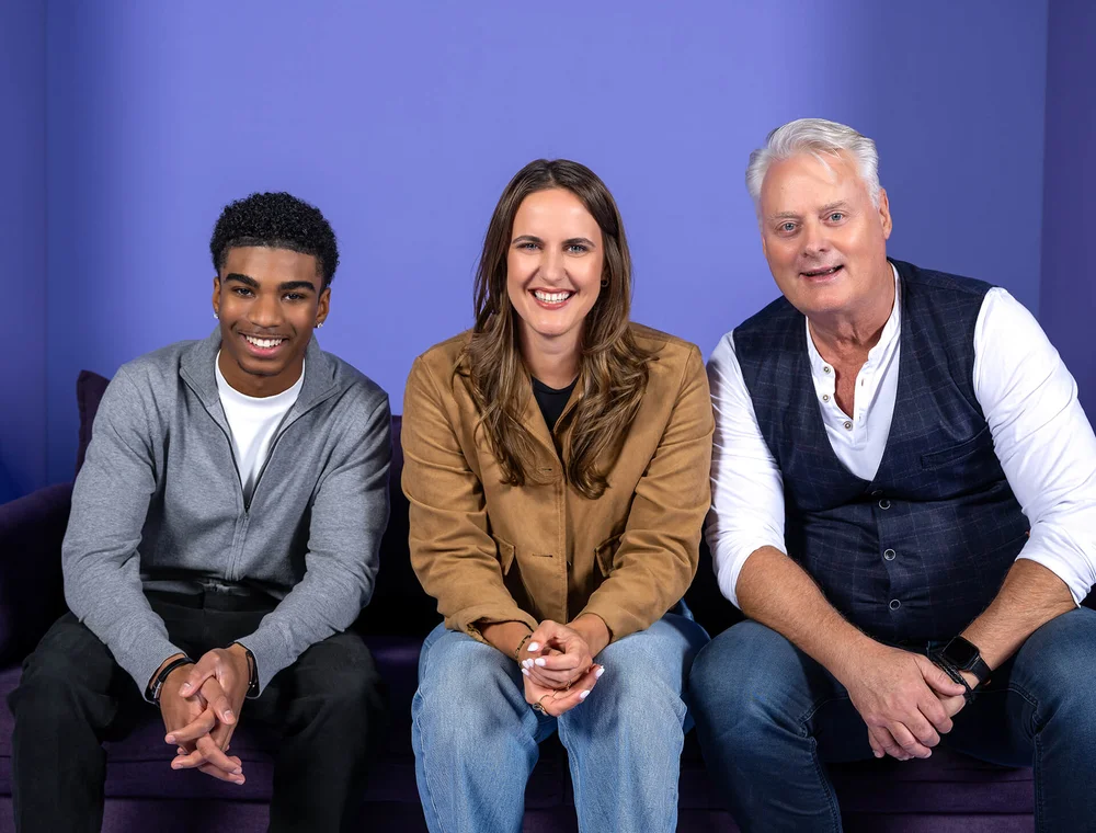 Drei Personen sitzen auf einem Sofa vor einem lila Hintergrund. Eine Frau sitzt in der Mitte zwischen zwei Männern. Alle lächeln freundlich in die Kamera. Die Szene wirkt entspannt und herzlich.