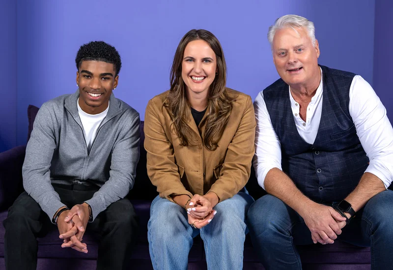 Drei Personen sitzen auf einem Sofa vor einem lila Hintergrund. Eine Frau sitzt in der Mitte zwischen zwei Männern. Alle lächeln freundlich in die Kamera. Die Szene wirkt entspannt und herzlich.