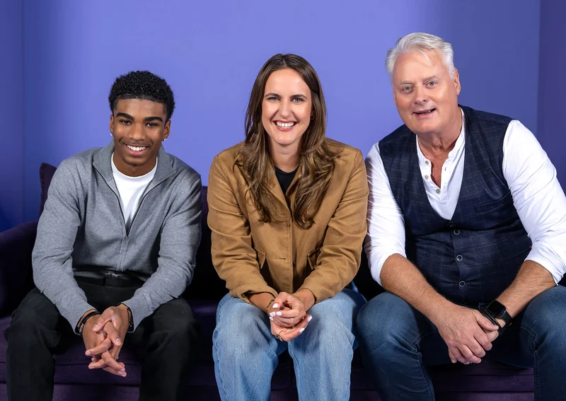 Drei Personen sitzen auf einem Sofa vor einem lila Hintergrund. Eine Frau sitzt in der Mitte zwischen zwei Männern. Alle lächeln freundlich in die Kamera. Die Szene wirkt entspannt und herzlich.