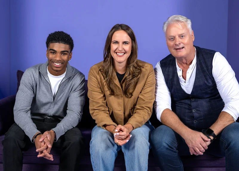 Drei Personen sitzen auf einem Sofa vor einem lila Hintergrund. Eine Frau sitzt in der Mitte zwischen zwei Männern. Alle lächeln freundlich in die Kamera. Die Szene wirkt entspannt und herzlich.