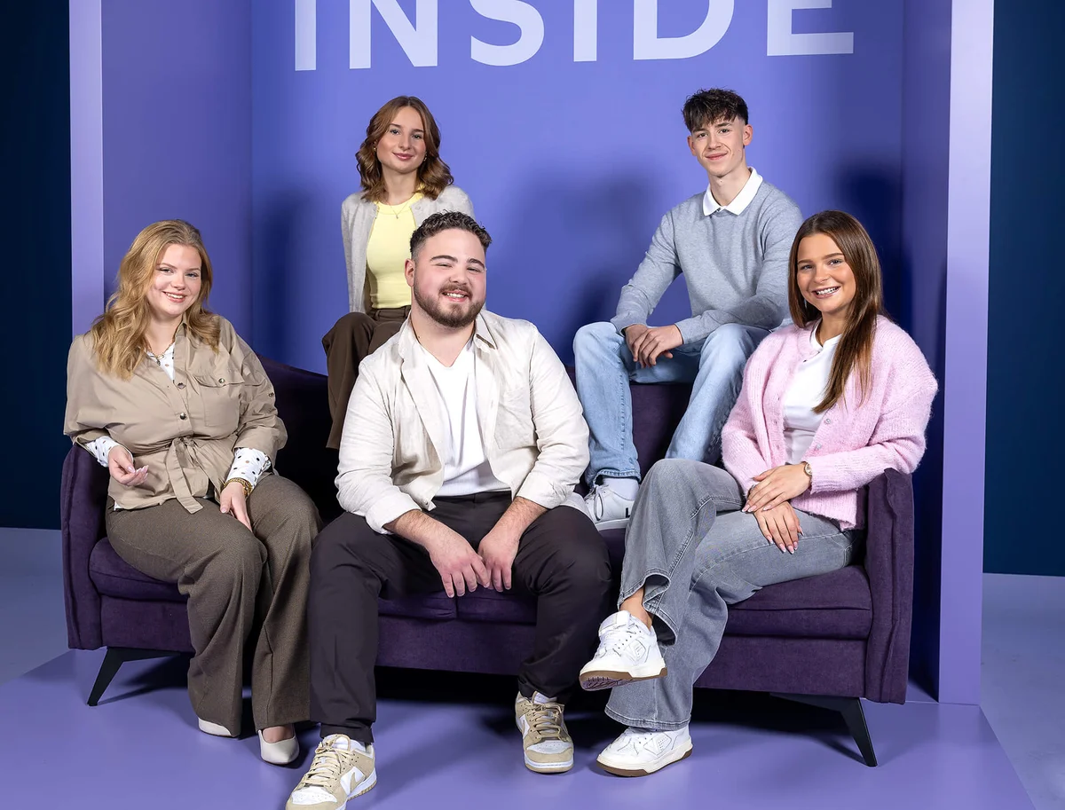 Fünf junge Erwachsene sitzen und stehen um ein lila Sofa vor einem violetten Hintergrund. Der Text "INSIDE" ist oben sichtbar. Alle lächeln in die Kamera, und ihre Kleidung ist modern und lässig.