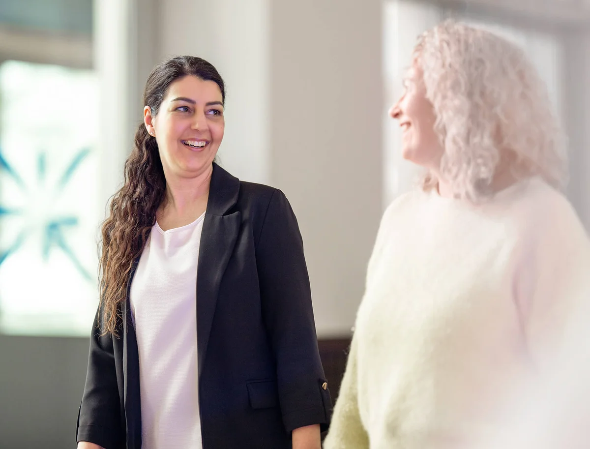 Zwei Frauen stehen in einem Raum und lächeln einander an. Eine Frau hat dunkles, langes Haar und trägt einen schwarzen Blazer, die andere hat lockiges, blondes Haar und trägt einen cremefarbenen Pullover. Sie wirken entspannt und freundlich.