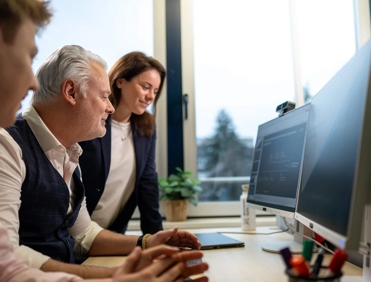 Zwei Männer und eine Frau betrachten konzentriert einen Computermonitor in einem Büro. Sie diskutieren offensichtlich über die angezeigten Informationen oder arbeiten gemeinsam an einem Projekt. Der Hintergrund zeigt ein Fenster mit unscharfem Ausblick nach draußen.