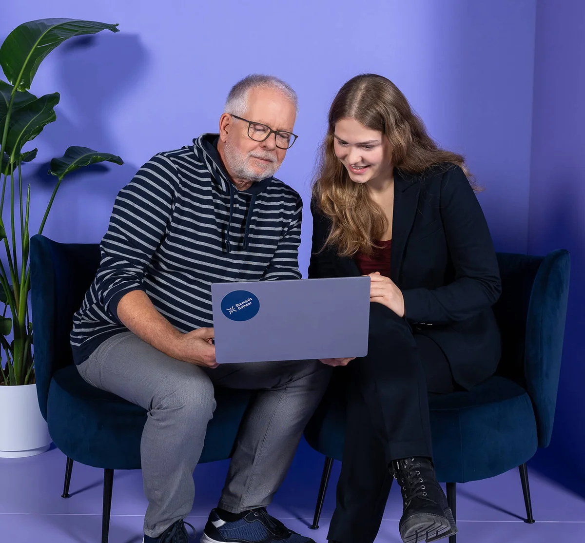 Ein älterer Mann und eine junge Frau sitzen nebeneinander auf einem Sofa und schauen gemeinsam auf einen Laptop. Beide wirken konzentriert und zufrieden. Im Hintergrund steht eine grüne Pflanze vor einer lila Wand.