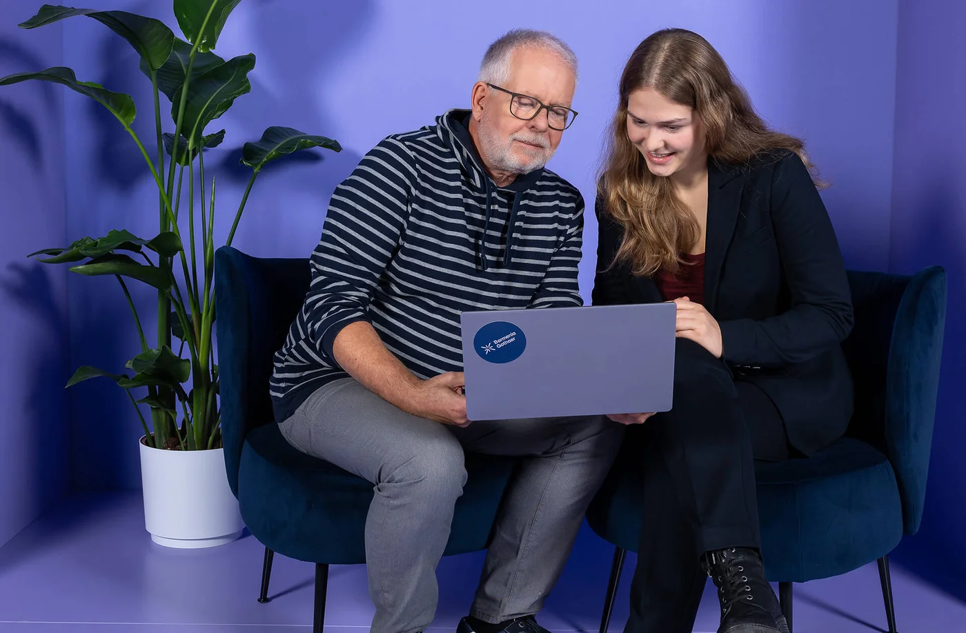 Ein älterer Mann und eine junge Frau sitzen nebeneinander auf einem Sofa und schauen gemeinsam auf einen Laptop. Beide wirken konzentriert und zufrieden. Im Hintergrund steht eine grüne Pflanze vor einer lila Wand.