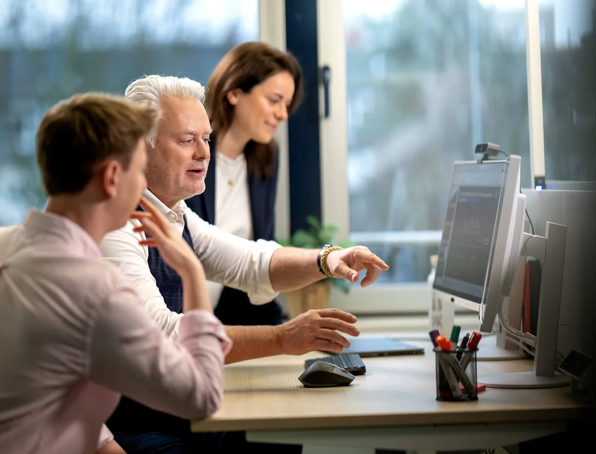 Eine Gruppe von drei Personen sitzt an einem Schreibtisch und schaut auf einen Computerbildschirm. Eine Person zeigt auf den Bildschirm, während die anderen beiden aufmerksam zuhören. Sie scheinen konzentriert und in eine Diskussion vertieft zu sein.