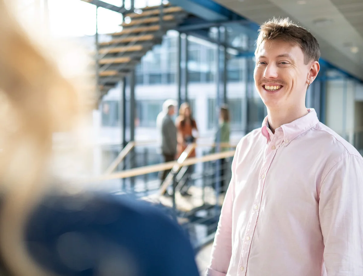 Ein junger Mann in einem rosa Hemd lächelt in einem modernen Gebäude. Im Hintergrund sind unscharf mehrere Personen und eine Treppe sichtbar.