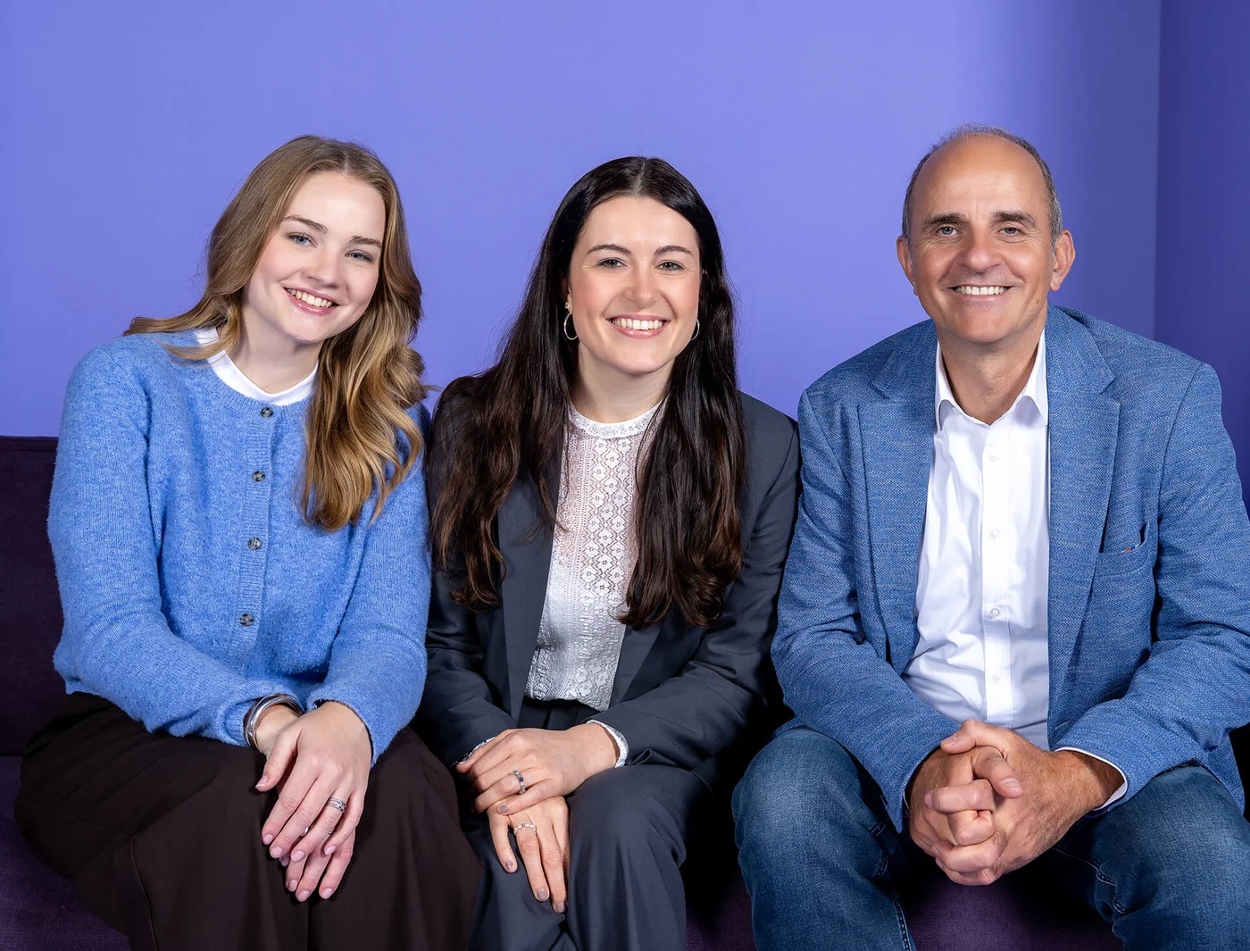 Drei Personen sitzen nebeneinander und lächeln in die Kamera. Links eine junge Frau mit langen blonden Haaren in blauer Jacke, in der Mitte eine Frau mit dunklen Haaren im Anzug, rechts ein Mann in blauem Sakko. Hintergrund ist ein lila Farbton.