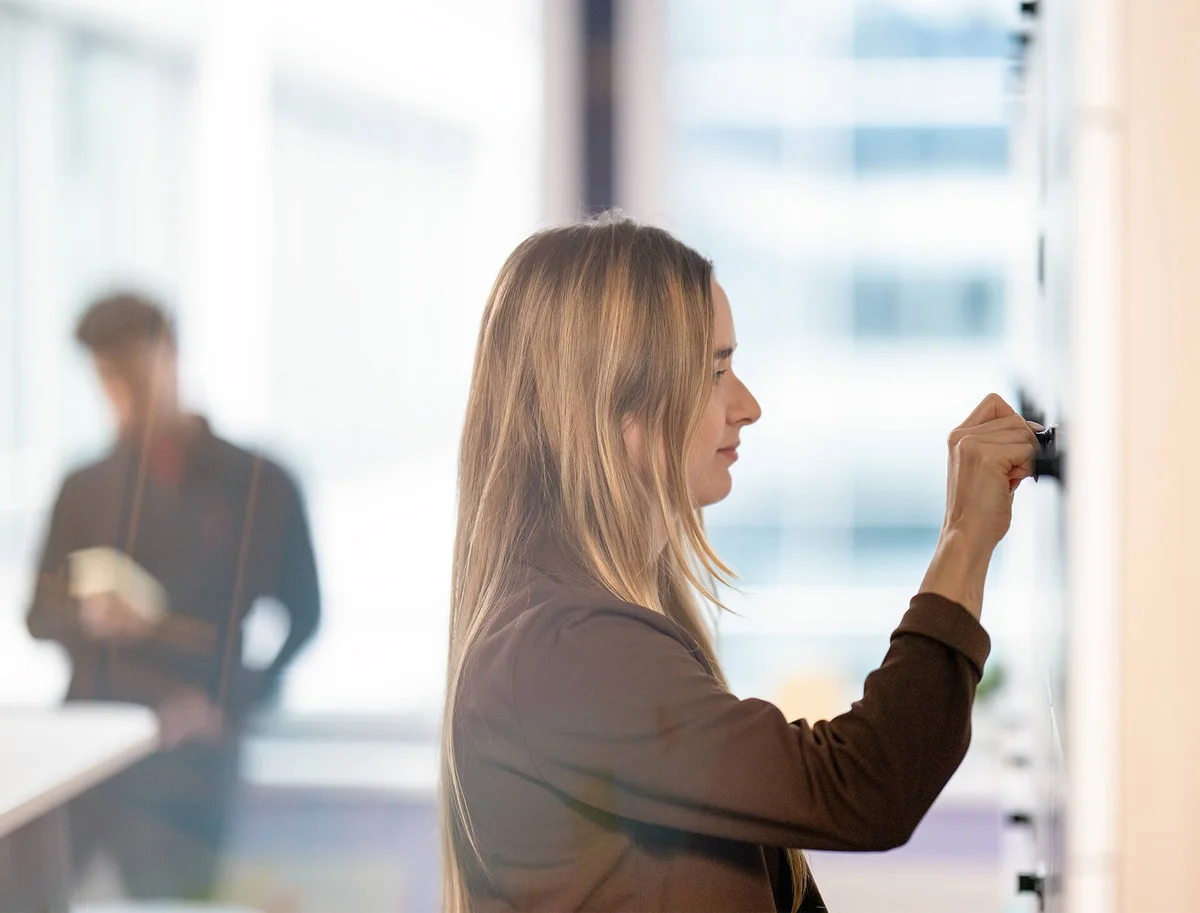 Eine Frau mit langen Haaren steht in einem Büro und nutzt ein Whiteboard. Im Hintergrund ist unscharf eine Person mit Notizblock zu sehen. Die Szene vermittelt einen professionellen Arbeitseindruck.