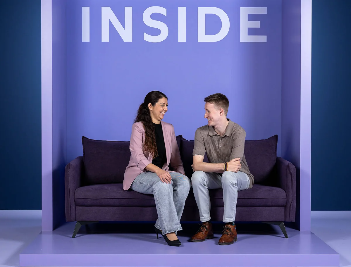 Ein Mann und eine Frau sitzen auf einem lila Sofa und lächeln sich an. Der Hintergrund ist ebenfalls lila, mit dem Wort "INSIDE" oben.