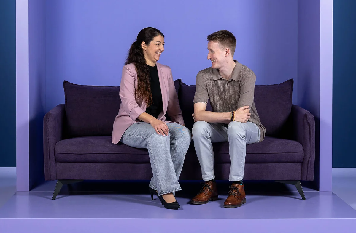 Ein Mann und eine Frau sitzen auf einem lila Sofa und lächeln sich an. Der Hintergrund ist ebenfalls lila, mit dem Wort "INSIDE" oben.