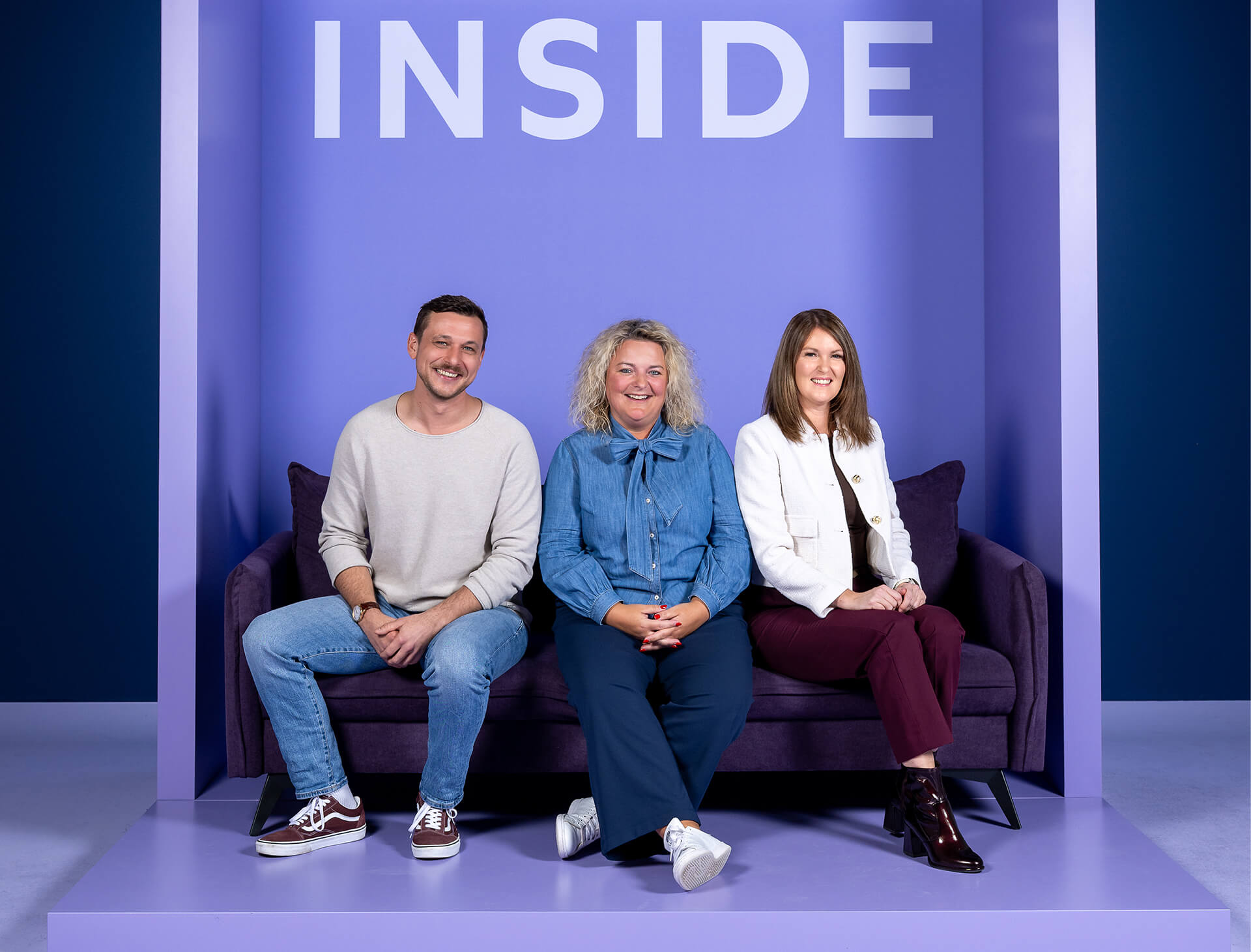 Ein Mann und zwei Frauen sitzen auf einem lila Sofa vor einem Hintergrund mit der Aufschrift "INSIDE". Alle lächeln in die Kamera.